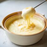 A piece of bread being lifted from a pot of hot, creamy melting pot cheese fondue, showing long, satisfying cheese pulls.