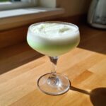 A bright green and white frothy mint white chocolate martini served in a coupe glass on a wooden counter.