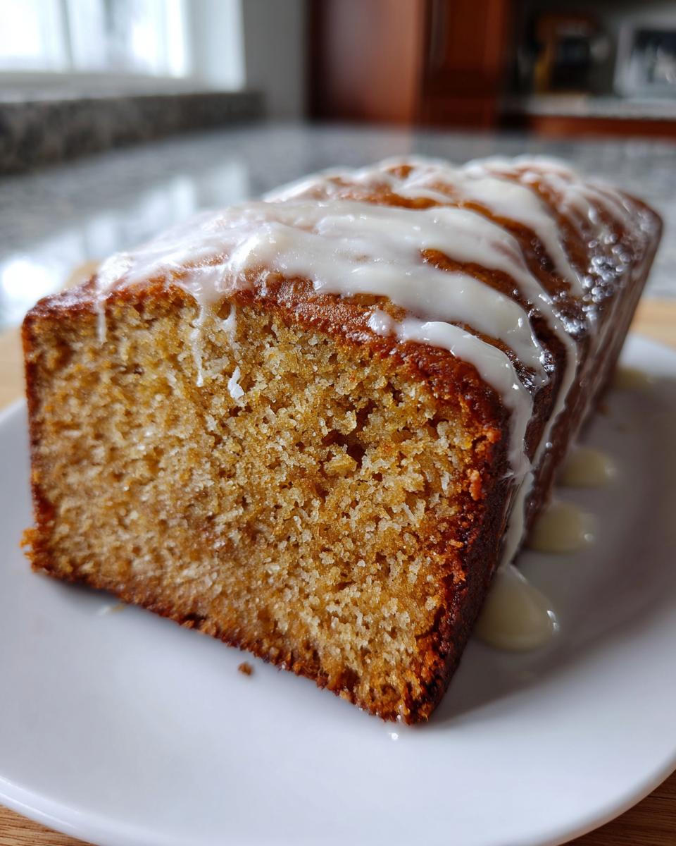 A close-up of a freshly baked, moist eggnog bread loaf drizzled with white icing on a white plate.