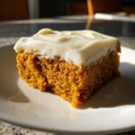 A close-up, sunlit slice of moist pumpkin bars topped with thick cream cheese frosting on a white plate.