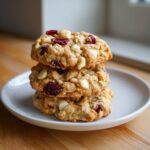 A stack of three chewy oatmeal cranberry white chocolate macadamia nut cookies on a white plate.