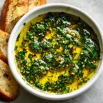 A white bowl filled with bright yellow olive oil bread dipping sauce featuring chopped parsley and garlic.