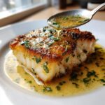 A perfectly pan seared Chilean sea bass fillet being drizzled with lemon butter sauce and fresh parsley.