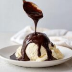 A spoonful of rich, dark chocolate sauce for ice cream is being poured over a scoop of vanilla ice cream.