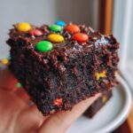 A close-up shot of a rich, dark chocolate protein cosmic brownie held in a hand, topped with colorful candy pieces.