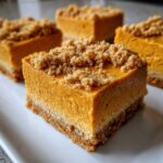 A close-up of a single, thick square of pumpkin cheesecake bars with a graham cracker crust and crumb topping.