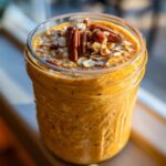 A close-up of creamy, orange pumpkin pie overnight oats topped with pecans and rolled oats in a glass jar.