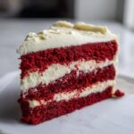 A perfect slice of red velvet cheesecake cake showing three vibrant red layers separated by thick cream cheese frosting.