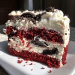 A decadent slice of red velvet oreo cheesecake showing layers of red cake, white cream filling with Oreo chunks, and topped with whipped cream.