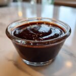 Close-up of thick, glossy homemade chocolate sauce served in a small clear glass bowl.