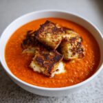 Close-up of vibrant roasted red pepper soup topped with crispy grilled cheese croutons.