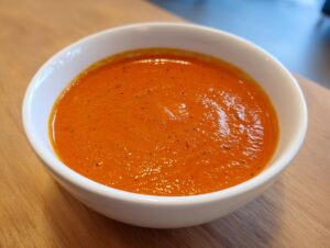 Close-up of a white bowl filled with vibrant orange roasted red pepper soup, seasoned with pepper flakes.