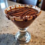A rich, brown salted caramel chocolate martini in a glass with a sugar/salt rim and chocolate drizzle.