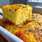 A square slice of golden Jiffy corn casserole being lifted from a white baking dish, topped with melted cheese and parsley.