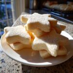 A stack of soft, thick, star-shaped sugar cookies dusted with fine sugar on a white plate.
