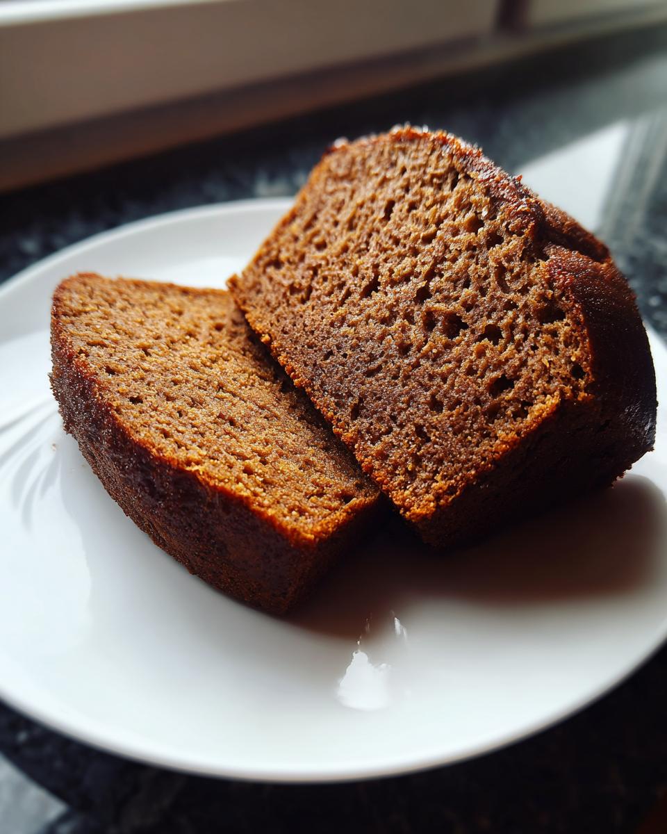 Two dark, moist slices of sourdough beer bread resting on a white plate near a window.