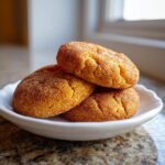 A close-up of three soft, golden pumpkin snickerdoodles generously coated in cinnamon sugar, stacked on a white plate.