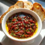 Close-up of sun-dried tomato olive oil bread dip with herbs, served with slices of crusty bread.