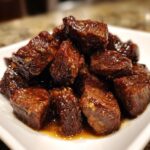 Close-up of tender crockpot steak bites piled high on a white plate, coated in a rich, glossy brown sauce.
