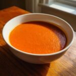 A close-up of a white bowl filled with vibrant orange tomato and roasted red pepper soup on a wooden table.
