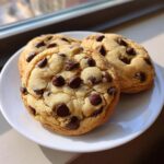 Three soft, thick vanilla pudding chocolate chip cookies piled on a white plate near a window.