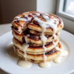 A tall stack of vegan cinnamon roll pancakes drizzled heavily with white icing.