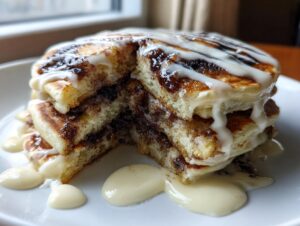 A stack of vegan cinnamon roll pancakes cut in half, drizzled heavily with white icing.