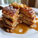 A thick stack of fluffy apple pancakes cut open, drizzled generously with caramel sauce and topped with cinnamon-spiced apple chunks.