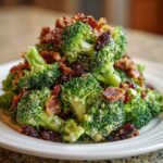 A close-up of a creamy broccoli salad topped with crispy bacon bits and dried raisins.