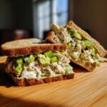 A hearty avocado chicken salad sandwich, cut in half, showing creamy chicken salad mixed with chunks of avocado and red onion on whole wheat bread.