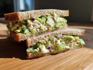 Two halves of an avocado chicken salad sandwich stacked on a wooden board, showing creamy filling with chicken, avocado, celery, and red onion.