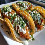 A plate of crispy, golden-brown baja fish tacos topped with a generous drizzle of white cream sauce and fresh cilantro.