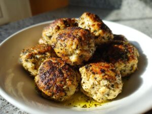 A pile of perfectly browned baked greek chicken meatballs seasoned with herbs, resting in a white bowl with olive oil.