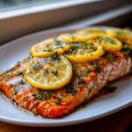 A close-up of juicy baked salmon fillets oven topped with fresh dill and overlapping lemon slices.