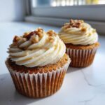 Two delicious banana walnut cupcakes topped with creamy frosting and chopped walnuts, sitting on a white marble surface.