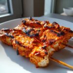 Close-up of grilled Bang Bang Chicken Skewers Moribyan, coated in a vibrant orange-red sauce and showing char marks, resting on a white plate.