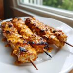 Three grilled bang bang chicken skewers generously coated in a creamy orange sauce, resting on a white plate.