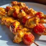 Close-up of grilled bang bang chicken skewers 12 tomatoes coated in a creamy orange sauce, served on a white plate.