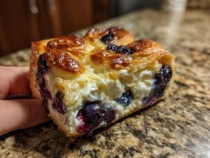 A close-up view of a square slice of blueberry cream cheese croissant bake, showing flaky pastry and creamy filling.