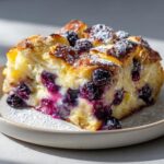 A close-up of a single serving of blueberry cream cheese croissant casserole, topped with powdered sugar.