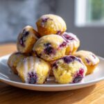 A stack of golden blueberry lemon pancake bites drizzled with a bright white lemon glaze, served on a white plate.