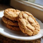 A stack of four golden brown butter sugar cookies, coated in sparkling sugar, sitting on a white plate near a window.