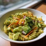A white bowl filled with creamy california pasta salad featuring rotini pasta, bright green broccoli florets, shredded carrots, and red onion.