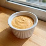 A close-up of creamy, orange-tinted cheeseburger egg roll sauce speckled with red spices in a white ramekin.