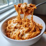 A spoonful of cheesy crockpot italian chicken being lifted from a white bowl, showing long, melty cheese strings.