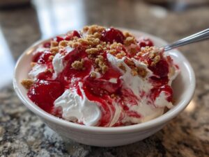 Close-up of a white bowl filled with creamy cherry cheesecake fluff, topped with cherry pie filling and graham cracker crumbs.