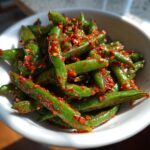 Close-up of vibrant green snap peas tossed heavily in bright red chili crisp, served in a white bowl.