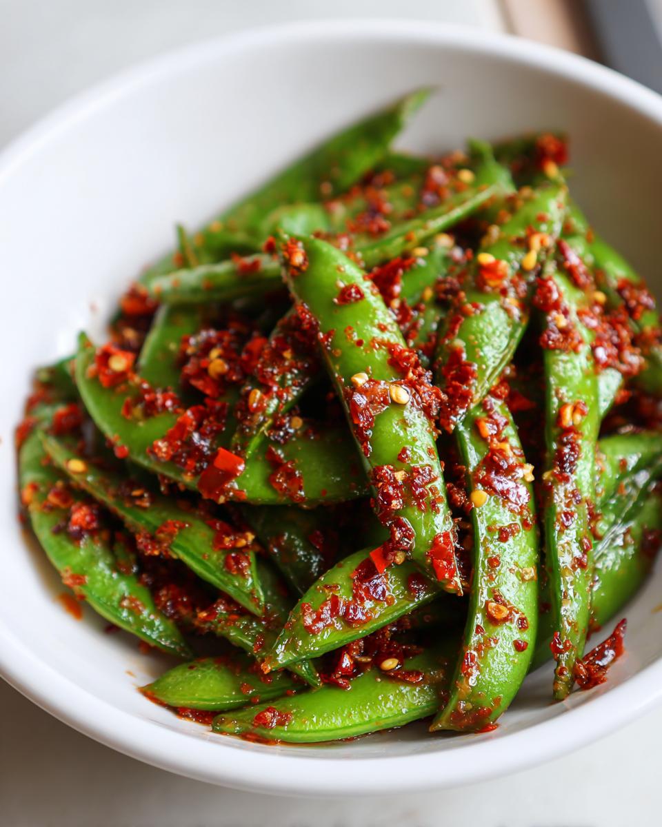 Close-up of bright green snap peas tossed generously with vibrant red chili crisp in a white bowl.