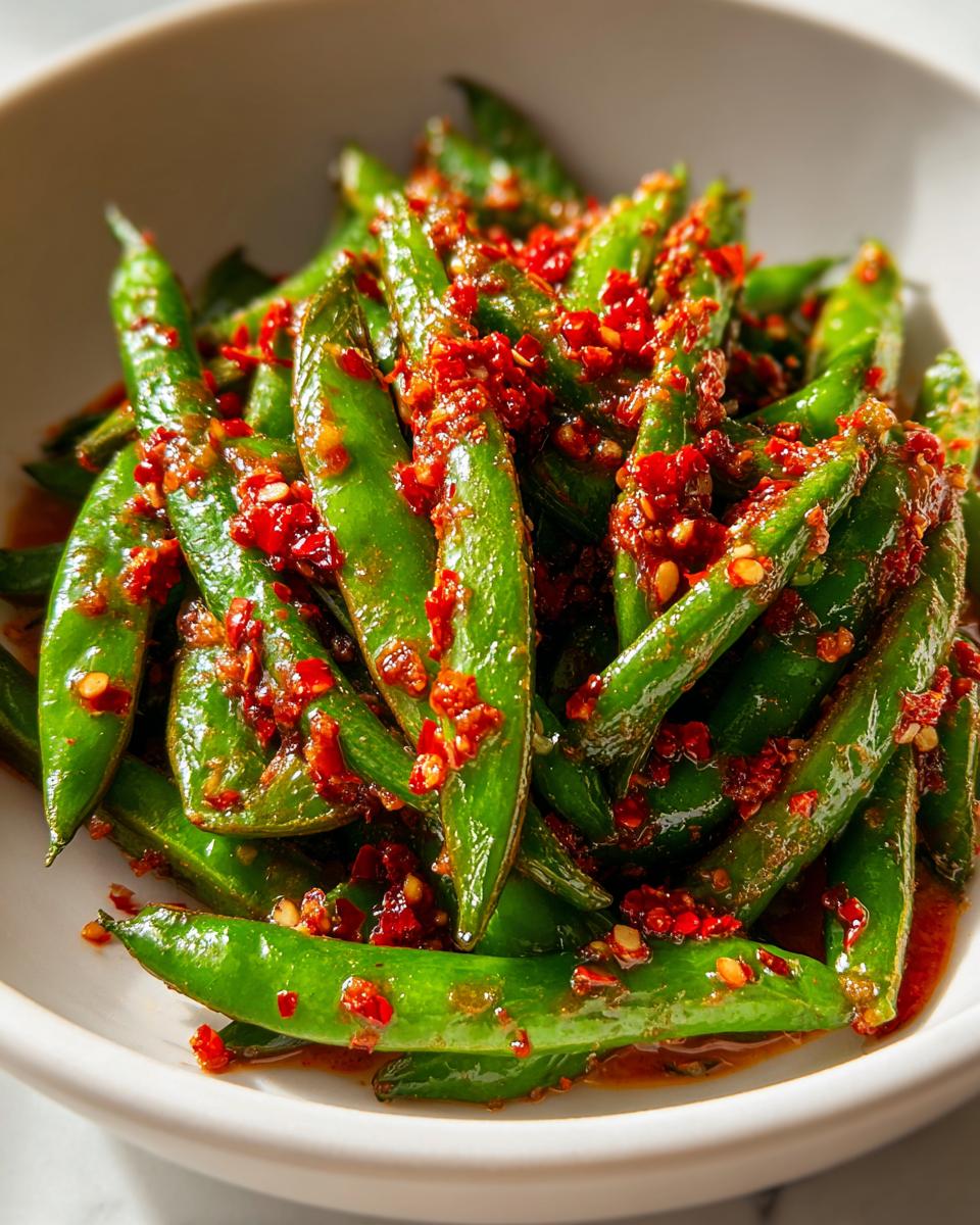 Close-up of bright green snap peas coated generously with red chili crisp, ready for a chili crisp snap pea salad.