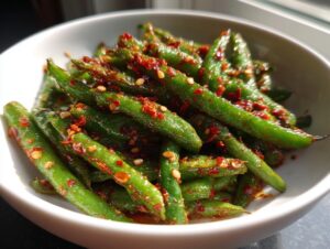 Close-up of bright green snap peas tossed generously with red chili crisp flakes and seeds, served in a white bowl.